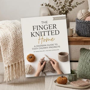 May include: A book titled "The Finger Knitted Home" with a subtitle "A Modern Guide to Cozy Chunky Projects" is displayed. The image features a cozy, homey scene with a chunky knit blanket, yarn balls, and a knitted basket. A pumpkin and a small knitted basket are also visible.