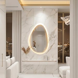 Puede incluir: Un espejo ovalado iluminado está montado en una pared de mármol. Una consola blanca con un jarrón y libros se encuentra debajo del espejo. La habitación cuenta con muebles blancos y una lámpara de araña.