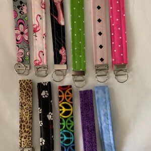 May include: Eleven fabric wristlet keychains arranged in two rows. Patterns include gray floral, pink flamingo, black high heels, green polka dot, light pink diamond, hot pink polka dot, leopard print, black paw print, rainbow peace sign, purple, and blue.