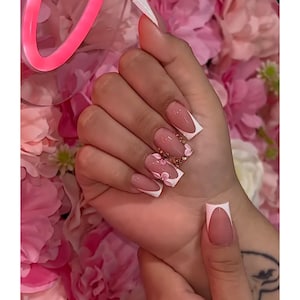 Puede incluir: Una mano con uñas largas y cuadradas. Las uñas presentan un diseño de manicura francesa con puntas blancas y una base nude. Algunas uñas están adornadas con diseños florales rosas y detalles de purpurina dorada.