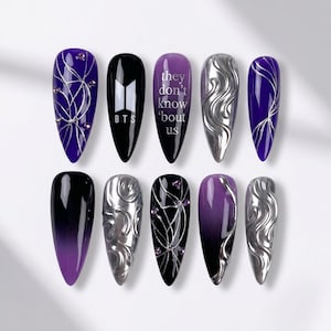 May include: A set of ten artificial nails in various designs. The nails feature a mix of colors including black, silver, and purple. Some nails have text that reads "they don't know 'bout us" and "BTS".