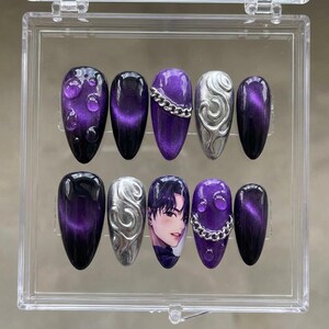 May include: A set of ten artificial nails in a clear plastic case. The nails feature a variety of designs, including a gradient of black to purple, silver chain accents, and a portrait of a person.