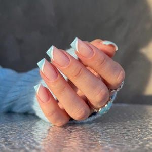 Op de afbeelding: Een hand met vierkante, lichtroze nagels met een French manicure. De nagelranden zijn lichtblauw met een witte diagonale accentlijn. Een zilveren ring met een turquoise hartvormige steen is zichtbaar.