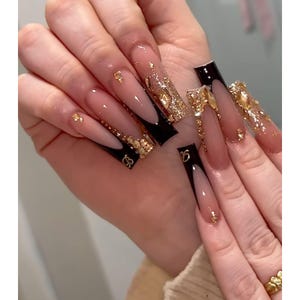 May include: Long, square-shaped acrylic nails with a French tip design. The nails feature a black and nude color scheme, adorned with gold glitter and small gold embellishments. Some nails have a gold letter design.