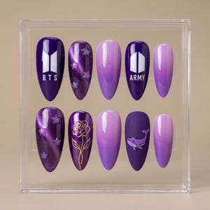 May include: A set of ten purple press-on nails in a clear display case. Designs include the BTS and ARMY logos, star patterns, a gold rose, and a whale. The nails have a glossy finish and a gradient effect.