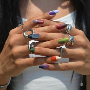 May include: Close-up of hands with long, oval-shaped nails painted in a variety of colors, including purple, red, green, and orange. Silver rings of various designs adorn the fingers. The nails feature a gradient effect, transitioning from one color to another.