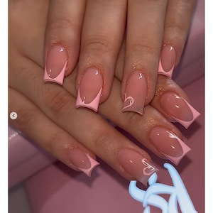 Pode incluir: Close-up de uma mão com unhas acrílicas longas e quadradas. As unhas são pintadas com uma base rosa claro e uma ponta francesa rosa brilhante. Algumas unhas apresentam um desenho de coração branco. As unhas são bem cuidadas e brilhantes.