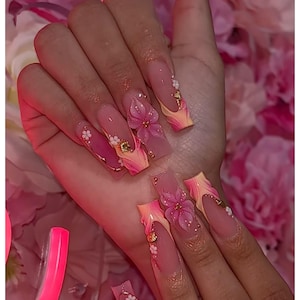 Könnte beinhalten: Lange, quadratische Acrylnägel mit einem rosa-gelben Farbverlauf. Jeder Nagel hat 3D-Blumendekorationen in Rosa, kleine weiße Blüten und goldene Akzente. Die Nägel sind mit kleinen, glitzernden Strasssteinen verziert.