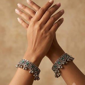 May include: A pair of silver-toned metal cuff bracelets featuring a rectangular geometric pattern with embedded circular green stones and small dangling silver-toned metal charms attached along the lower edge of each bracelet band.