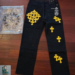 May include: Black denim jeans featuring a large cluster of yellow cross patches on the left leg and scattered yellow cross patches on the right leg. A white graphic print of a cross and the text CHROME HEARTS is visible on the plastic packaging.
