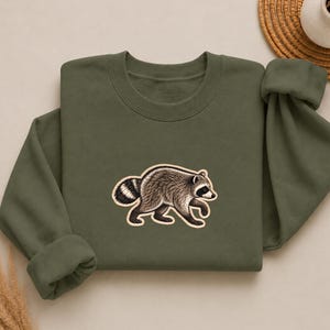 May include: A military green crewneck sweatshirt featuring a detailed, centered graphic illustration of a raccoon in shades of black, white, and gray. The long-sleeved pullover has a ribbed neckline, cuffs, and hem, showcasing a clean, solid design.