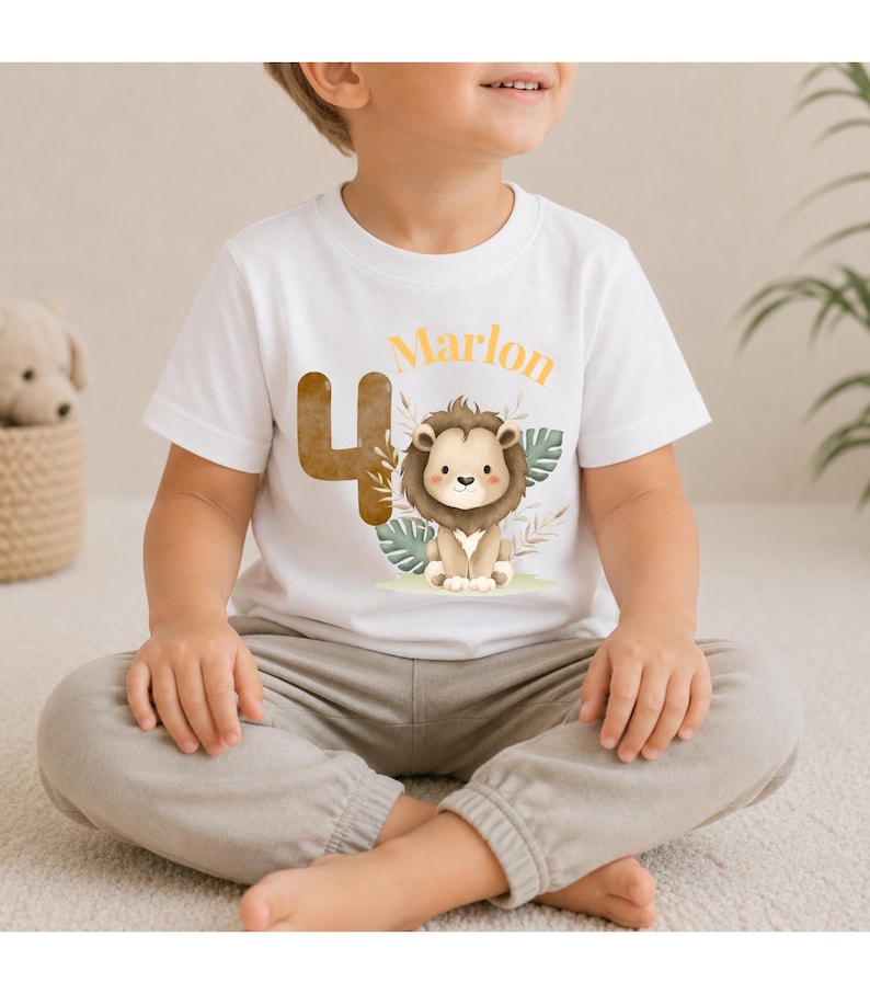 May include: A white short-sleeved t-shirt featuring a graphic print of a brown number 4, a cartoon lion, and the name Marlon in orange text. The design includes green tropical leaves and beige foliage accents centered on the chest area.