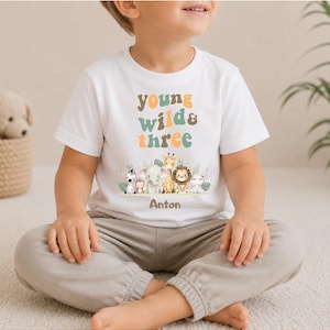 Puede incluir: Una camiseta blanca de manga corta con el texto "young wild & three" en tonos naranja y verde apagados. Debajo del texto hay una ilustraci&oacute;n gr&aacute;fica de una cebra, un mono, un elefante, una jirafa, un le&oacute;n y un hipop&oacute;tamo, con el nombre "Anton" impreso debajo.