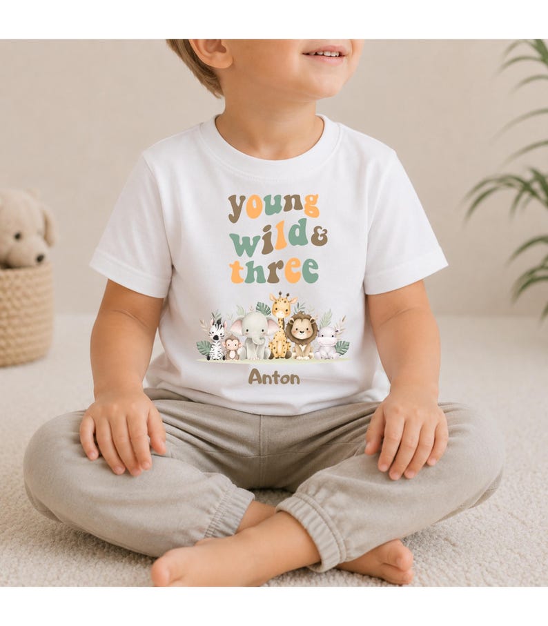 Puede incluir: Una camiseta blanca de manga corta con el texto "young wild & three" en tonos naranja y verde apagados. Debajo del texto hay una ilustraci&oacute;n gr&aacute;fica de una cebra, un mono, un elefante, una jirafa, un le&oacute;n y un hipop&oacute;tamo, con el nombre "Anton" impreso debajo.