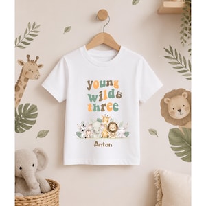 Puede incluir: Una camiseta blanca de manga corta con el texto "young wild & three" en letras multicolores sobre una ilustraci&oacute;n gr&aacute;fica de una cebra, un mono, un elefante, una jirafa, un le&oacute;n y un hipop&oacute;tamo, con el nombre "Anton" impreso en marr&oacute;n debajo.