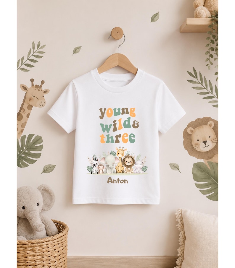 Puede incluir: Una camiseta blanca de manga corta con el texto "young wild & three" en letras multicolores sobre una ilustraci&oacute;n gr&aacute;fica de una cebra, un mono, un elefante, una jirafa, un le&oacute;n y un hipop&oacute;tamo, con el nombre "Anton" impreso en marr&oacute;n debajo.