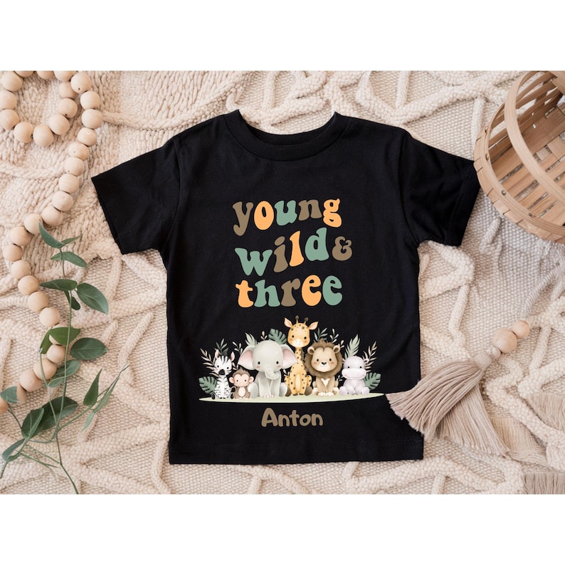 Puede incluir: Una camiseta negra de manga corta con el texto "young wild & three" en naranja y verde, junto a un gr&aacute;fico de una cebra, un mono, un elefante, una jirafa, un le&oacute;n y un hipop&oacute;tamo. El nombre "Anton" aparece impreso en marr&oacute;n bajo los animales.