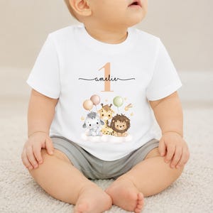 May include: White short-sleeve t-shirt featuring a whimsical design for a first birthday. The design includes the number "1", the name "amelie", and illustrations of a lion, giraffe, elephant, and balloons. The shirt is worn by a baby.