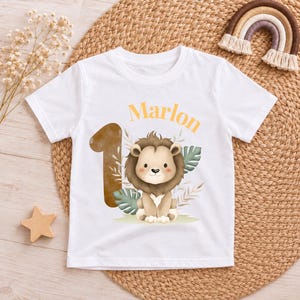 May include: A white short-sleeved t-shirt featuring a graphic print of a brown lion, a large number one, and the name Marlon in orange text. The design includes green tropical leaves and beige foliage accents centered on the front of the shirt.