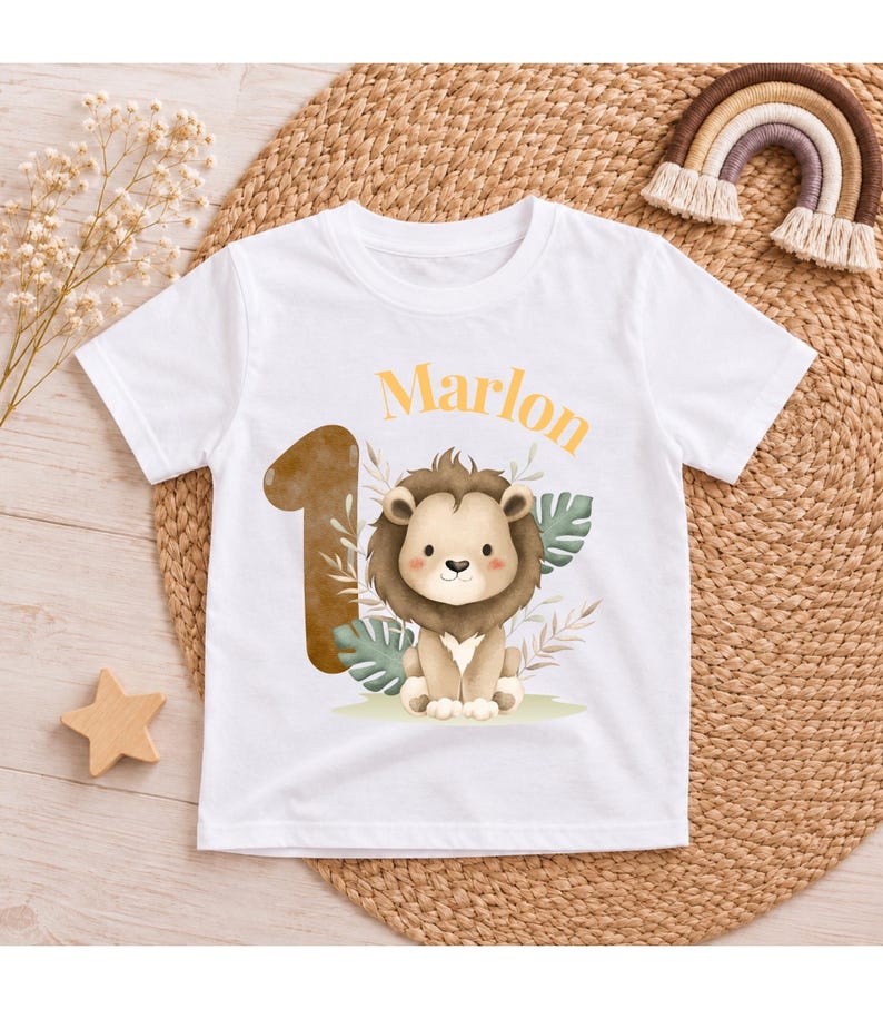 May include: A white short-sleeved t-shirt featuring a graphic print of a brown lion, a large number one, and the name Marlon in orange text. The design includes green tropical leaves and beige foliage accents centered on the front of the shirt.