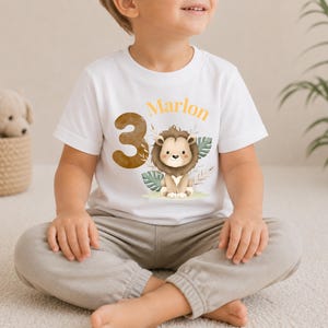 May include: A white short-sleeved t-shirt featuring a graphic of a cartoon lion, the number 3 in a brown textured font, and the name Marlon in orange text. The design includes watercolor-style green leaves and beige foliage accents.