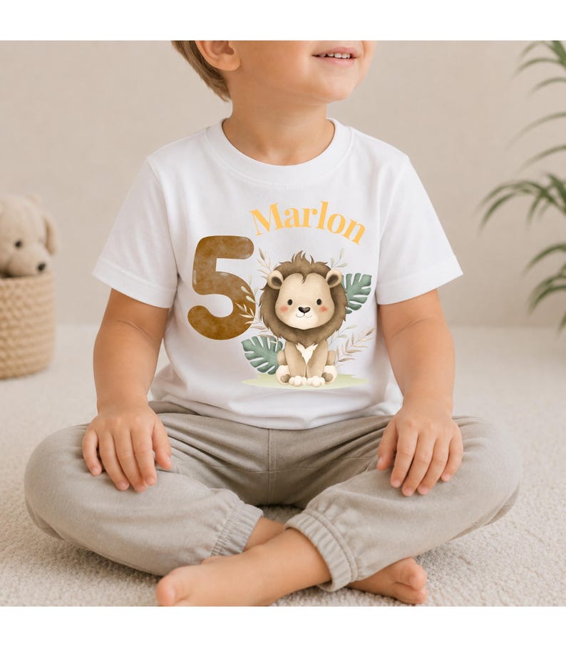 May include: A white short-sleeved t-shirt featuring a large brown number 5, a cartoon lion illustration, and the name Marlon in orange text. The design includes watercolor-style tropical leaves and foliage surrounding the lion character.