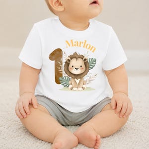 May include: A white short-sleeved t-shirt featuring a graphic of a brown lion, a large brown number one, and green tropical leaves. The name Marlon is printed in orange above the lion. The shirt is paired with plain grey shorts.