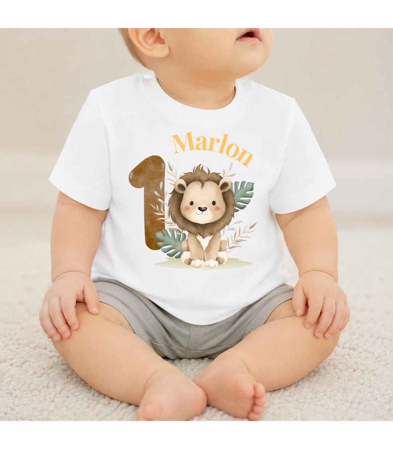 May include: A white short-sleeved t-shirt featuring a graphic of a brown lion, a large brown number one, and green tropical leaves. The name Marlon is printed in orange above the lion. The shirt is paired with plain grey shorts.