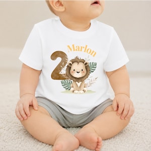 May include: A white short-sleeved t-shirt featuring a large brown number two, a watercolor illustration of a lion, green tropical leaves, and the name Marlon printed in orange text across the front of the garment.