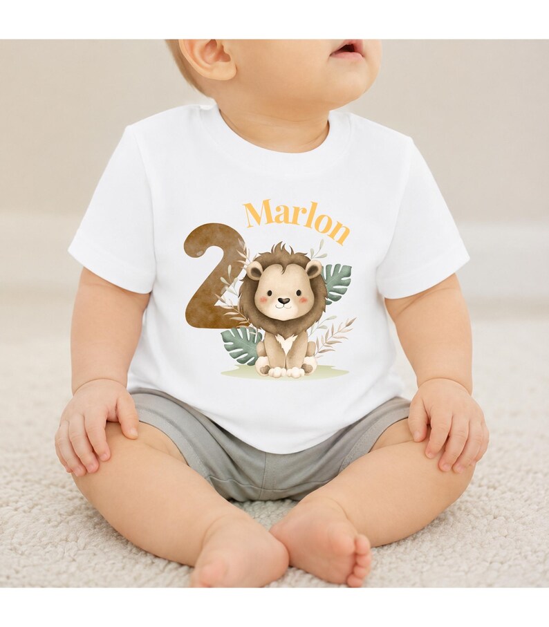 May include: A white short-sleeved t-shirt featuring a large brown number two, a watercolor illustration of a lion, green tropical leaves, and the name Marlon printed in orange text across the front of the garment.