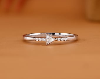 Op de afbeelding: Een zilverkleurige ring met een centrale driehoekig geslepen heldere steen, geflankeerd door drie kleine rond geslepen heldere steentjes aan weerszijden van de band. De ring heeft een gepolijste metalen afwerking en de tekst AVIRA FINE JEWELRY.