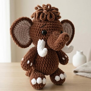 May include: A handmade crochet mammoth toy in shades of brown with white tusks and accents. The plush animal has large ears, a trunk, and a tuft of hair on its head. The toy is standing on a wooden surface.
