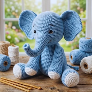 Puede incluir: Un elefante azul claro tejido a ganchillo con detalles blancos en los pies. El animal de peluche hecho a mano tiene orejas grandes, una trompa y una cara sonriente. Ganchos de ganchillo y ovillos de lana de varios colores en el fondo.
