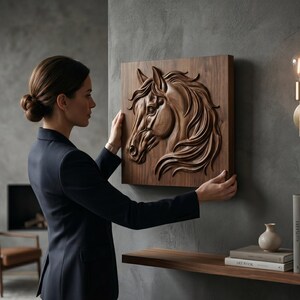 May include: A detailed, wooden relief carving of a horse's head, mounted on a square wooden panel. The artwork is a warm brown color and is being held by a person in a dark blue blazer. The horse's mane and facial features are intricately carved.