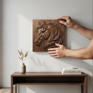 May include: A detailed, wooden relief carving of a horse's head is being mounted on a white wall. The artwork is a square, brown panel with the horse's profile in high relief. A minimalist interior setting includes a wooden table and a vase.