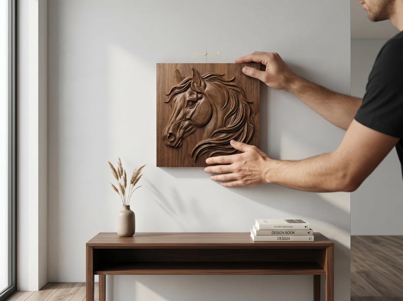 May include: A detailed, wooden relief carving of a horse's head is being mounted on a white wall. The artwork is a square, brown panel with the horse's profile in high relief. A minimalist interior setting includes a wooden table and a vase.