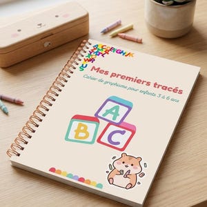Peut inclure: Un cahier à spirales avec le texte "Mes premiers tracés" et des illustrations colorées. La couverture présente des lettres de l'alphabet, des blocs ABC et un mignon hamster. Des crayons et une boîte en bois sont également visibles.
