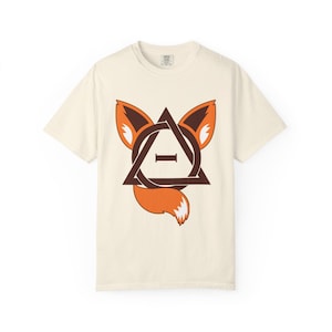 Könnte beinhalten: Cremefarbenes T-Shirt mit einem stilisierten Fuchsmotiv. Die Grafik zeigt ein braunes Dreieck, einen Kreis und den Buchstaben "I" in der Mitte, mit Fuchsohren und -schwanz in Orange und Braun. Ein lässiges Kleidungsstück.