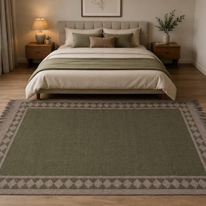 May include: A rectangular olive green rug with a geometric border and fringe detail. The rug is placed in front of a bed with neutral-colored bedding. The room has a warm, inviting atmosphere.