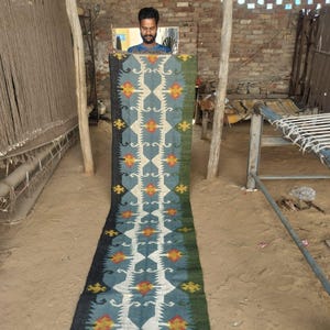 May include: A long, rectangular rug with a geometric pattern in shades of blue, white, yellow, and red. The rug has a dark blue border and a green edge. The rug is displayed vertically, showcasing its design and texture.