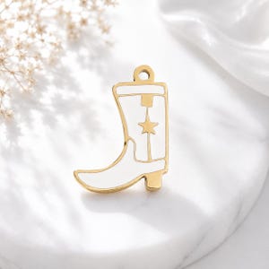 Könnte beinhalten: Ein goldfarbener Metallanhänger in Form eines Cowboystiefels mit weißer Emaille-Einlage und einem kleinen sternförmigen Ausschnitt am Schaft. Der Anhänger verfügt über eine kreisförmige Öse an der Oberseite zur Befestigung und zeichnet sich durch klare Linien und eine polierte Metalloberfläche aus.