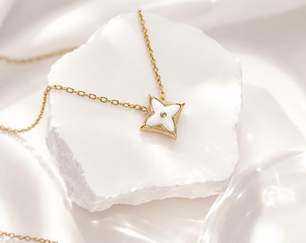 Pode incluir: Um colar dourado com um pingente de flor de quatro pétalas. O pingente tem um centro de esmalte branco com uma pequena pedra preciosa transparente. O colar é exibido em uma superfície branca texturizada.
