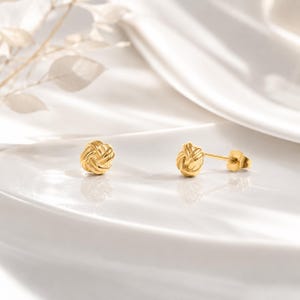 Peut inclure: Deux boucles d'oreilles dorées en forme de nœud tressé. Elles sont présentées sur une surface blanche, avec une boucle d'oreille montrant la tige et le fermoir. Ces boucles d'oreilles sont un bijou classique.