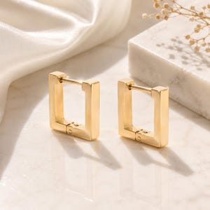 Könnte beinhalten: Ein Paar goldfarbene quadratische Creolen. Die Ohrringe haben eine rechteckige Form mit einem Klappverschluss. Die Ohrringe werden auf einer Marmoroberfläche mit weißem Satinstoff im Hintergrund präsentiert.