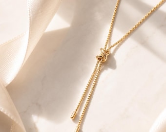 Pode incluir: Um colar tipo lariat em tom dourado com uma delicada corrente de elos quadrados. A corrente é atada num nó central, com duas pontas pendentes terminadas em pequenas tampas cilíndricas lisas. O metal tem uma superfície polida e brilhante.