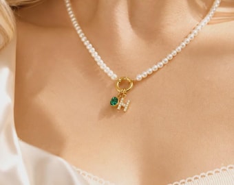 Pode incluir: Um colar de pérolas brancas com um fecho circular dourado. Pendurados no fecho estão dois berloques: uma pequena pedra redonda facetada em verde esmeralda e uma letra H dourada adornada com pequenos cristais claros e brilhantes.