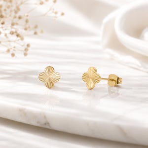 Peut inclure: Une paire de boucles d'oreilles clous dorées en forme de trèfle. Chaque boucle d'oreille présente un motif texturé et rayonnant. Les boucles d'oreilles sont présentées sur une surface en marbre blanc, avec un éclairage doux et un accent floral flou en arrière-plan.