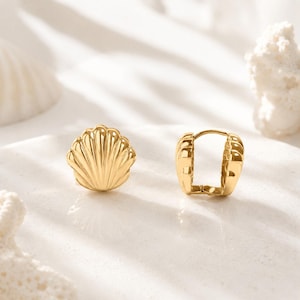 Puede incluir: Un par de pendientes dorados. Uno tiene forma de concha y el otro es una pequeña argolla. Los pendientes están sobre una superficie blanca con una concha y coral de fondo. Los pendientes son probablemente de metal.