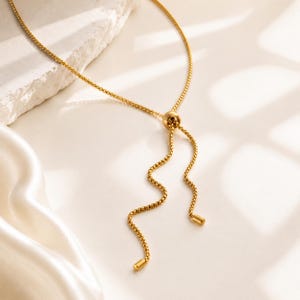 Pu&ograve; includere: Una collana a catena veneziana color oro con un meccanismo a perla scorrevole che consente di regolare la lunghezza. La catena termina con due fili pendenti, ciascuno rifinito con un piccolo tappo cilindrico in metallo dorato sulla punta.