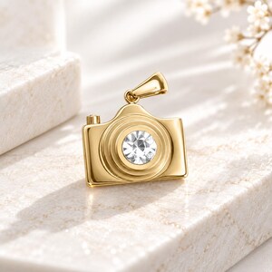 May include: A gold-toned metal pendant shaped like a vintage camera, featuring a central circular clear gemstone set into the lens area and a small top loop for attachment to a chain or cord, showcasing a polished and reflective metallic finish.